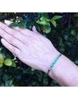 2.40 ct. emerald and diamond 14K yellow gold bracelet on a woman's wrist against green leaves.