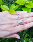 Hand displaying a four-leaf pendant with an emerald gemstone against a green leafy background.