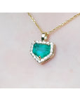 Gold necklace with a heart-shaped emerald pendant on a light pink background.