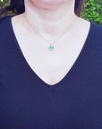 A woman wearing a dark blue top with a necklace featuring an emerald pendant against a blurred green background.