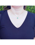A woman wearing a dark blue top with a necklace featuring an emerald pendant against a blurred green background.