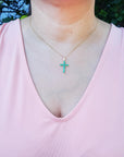 A woman wearing a pink top with a genuine emerald cross necklace against a blurred green background.