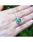 18k gold heart pendant with a bluish-green emerald haloed by diamonds, worn on a hand.