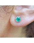 Round cut emerald stud earring in 14K white gold with a hexagonal diamond halo on an ear.