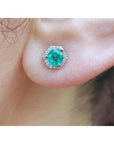Round cut emerald stud earring in 14K white gold with hexagonal diamond halo on an ear.