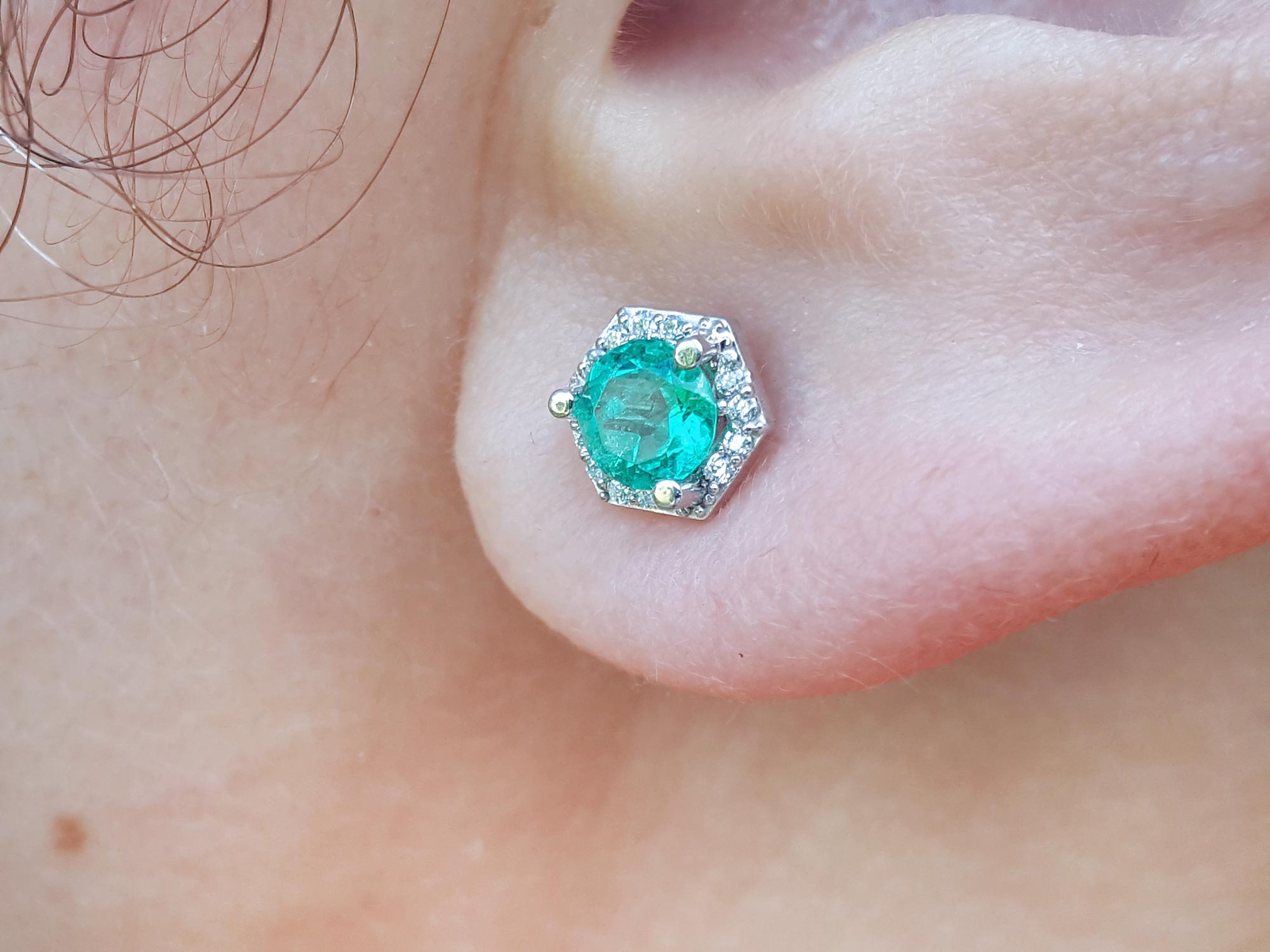 A geometri hexagonal emerald stud earrings with halo diamond worn on an earlobe close-up view.