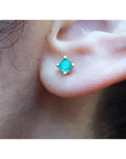 Close-up of an ear wearing a emerald stud earring with a blurred natural background.