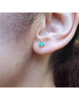 Close-up of an ear wearing a emerald stud earring with a blurred natural background.