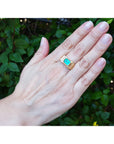 Hand wearing a ring with a natural colombian emerald against a green leafy background.