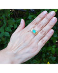 Hand wearing a ring with a natural colombian emerald against a green leafy background.