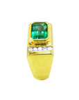 Gold ring for men with a bezel set natural emerald and smaller clear diamonds on a side profile position.