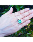 Cushion cut Colombian emerald necklace with halo diamonds on an 18K yellow gold chain, held in a hand.