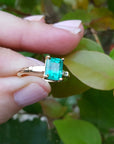 Gold ring with a colombian emerald held between fingers against a leafy background.