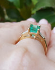 A yellow gold emerald ring with side diamonds on a hand against a blurred green leafy background.