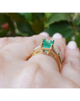 A yellow gold emerald ring with side diamonds on a hand against a blurred green leafy background.
