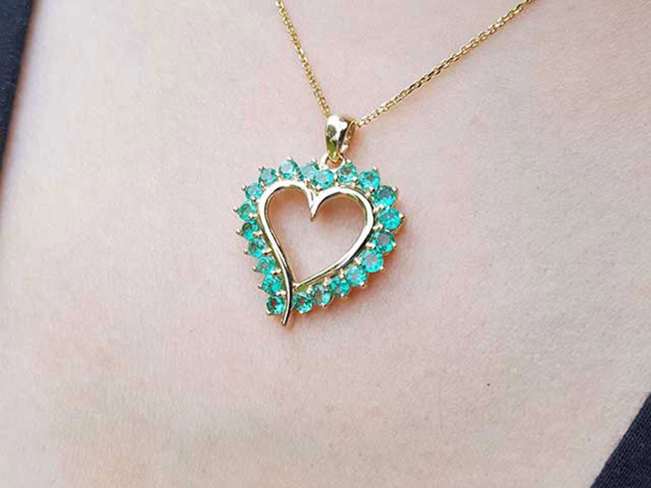 Heart-shaped pendant with emeralds gemstones on a gold chain against a light skin background.