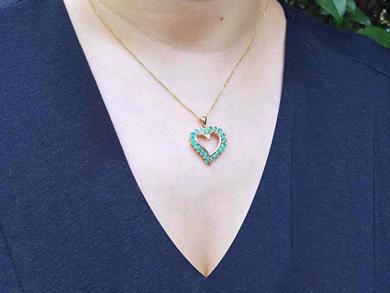 Heart-shaped pendant necklace with round cut emeralds on a person wearing a blue top.
