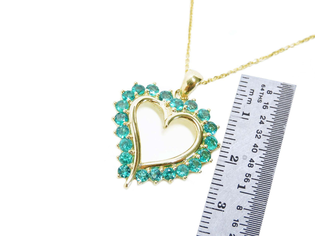 Gold heart-shaped pendant with round emerald stones next to a ruler for scale on a white background.