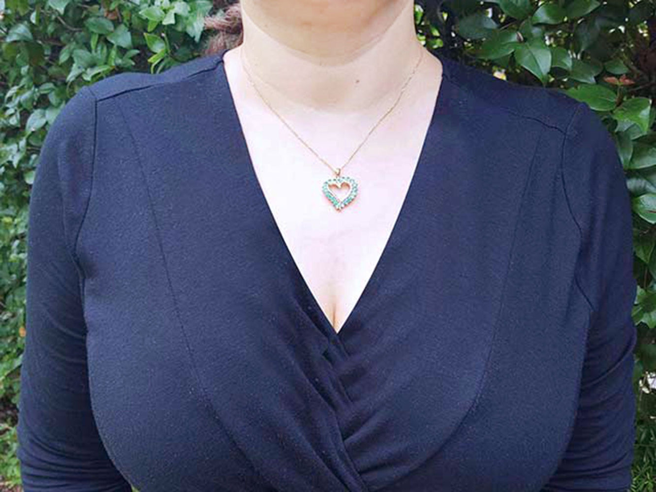 A woman wearing a navy-blue top with a natural emerald heart-shaped pendant necklace against a green leafy background.