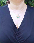 A woman wearing a navy-blue top with a natural emerald heart-shaped pendant necklace against a green leafy background.