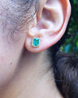 Close-up front view of an ear wearing a 14k gold emerald sud earring with a blurred green background