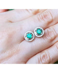 Halo emerald stud earrings with deep bluish-green emeralds encircled by diamonds, set in 14K white gold.