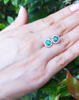 Hand displaying a pair of emerald stud earrings with a green leafy background