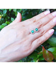 Halo emerald stud earrings with deep bluish-green 1.06 ct. emeralds encircled by diamonds on a hand.