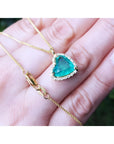 Tilted heart emerald necklace with halo diamonds in 18K yellow gold, shown on a hand.