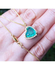 Tilted heart emerald necklace with halo diamonds in 18K yellow gold, shown on a hand.