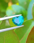 1.26 ct heart cut Colombian emerald, vibrant bluish-green, held by tweezers against green leaves.