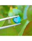 1.26 ct heart cut Colombian emerald, vibrant bluish-green, held by tweezers against green leaves.