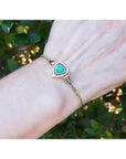 14K yellow gold bracelet with a heart-shaped emerald surrounded by diamonds on a woman's wrist.