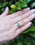 Hand wearing a heart cut emerald ring with a green leafy background.