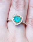 Heart-shaped emerald ring with halo diamond on a finger with a blurred green background.