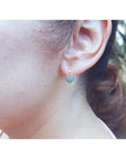 14K yellow gold heart earrings with brilliant cut Colombian emeralds, worn on an ear.