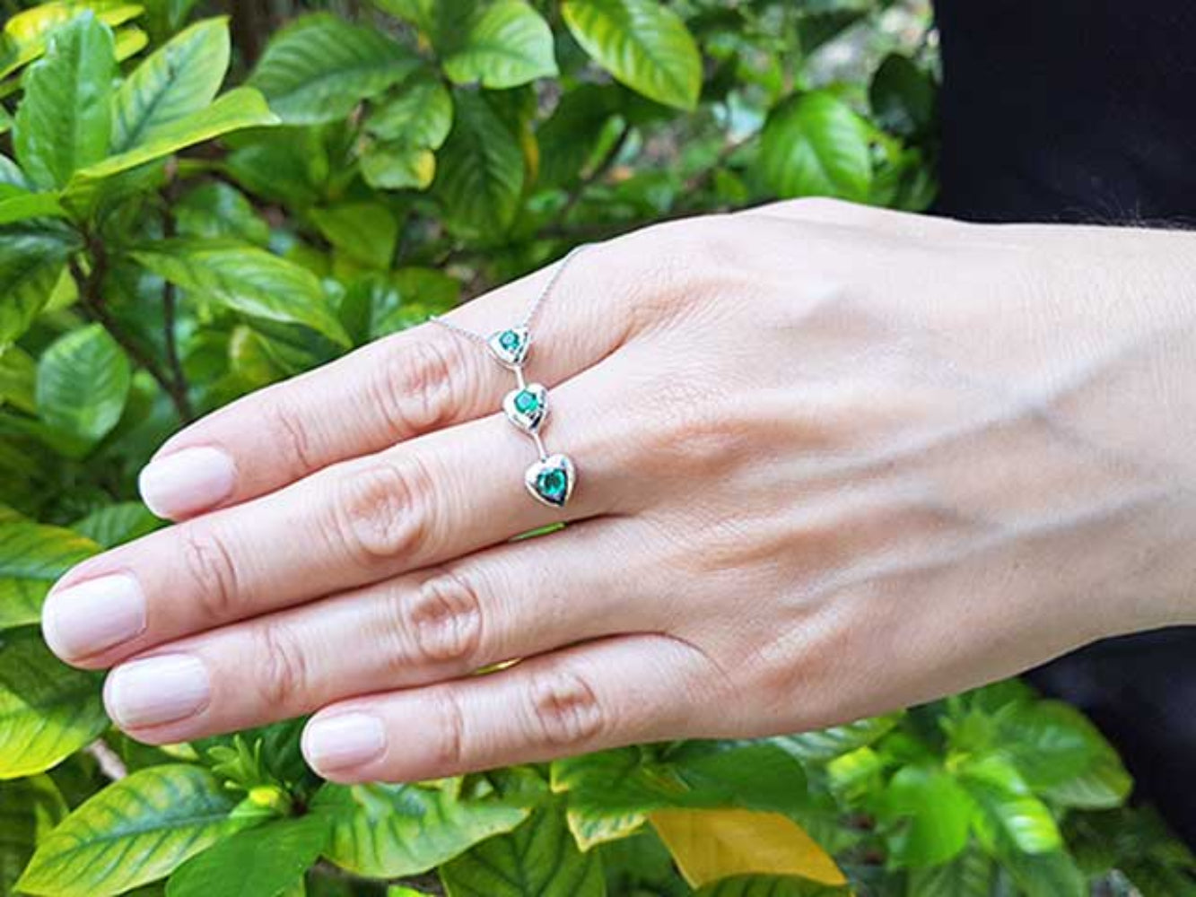Hand displaying a three links heart shaped journey pendant with round cut emeralds against a green leafy background.