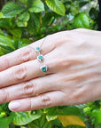 Hand displaying a three links heart shaped journey pendant with round cut emeralds against a green leafy background.