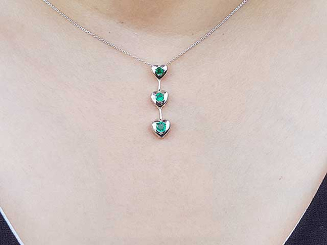 White gold necklace with heart-shaped emerald pendants on a person wearing a light-colored top.