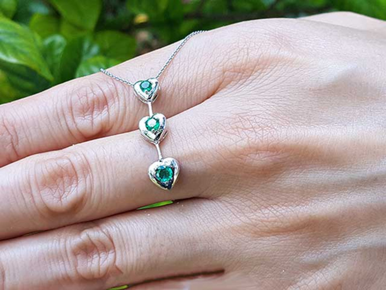 White gold necklace with heart-shaped emerald stones worn on a hand against a natural background.