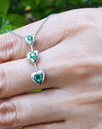 White gold necklace with heart-shaped emerald stones worn on a hand against a natural background.