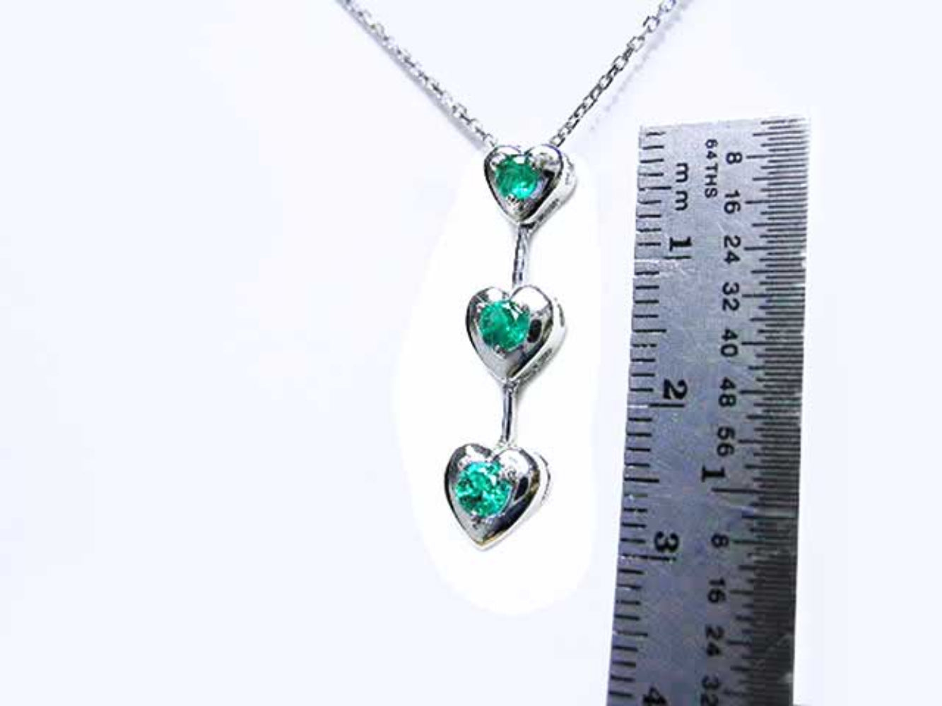 Necklace with heart-shaped pendants featuring natural emeralds, next to a ruler for scale on a white background.