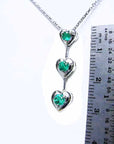 Necklace with heart-shaped pendants featuring natural emeralds, next to a ruler for scale on a white background.