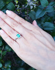 Hand wearing an emerald engagement ring against a leafy background.