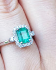 An inexpensive emerald engagement ring with a halo diamond on a finger.