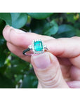 14K white gold emerald-cut emerald ring with diamond halo, held against green leaves.