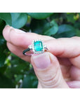 14K white gold emerald-cut emerald ring with diamond halo, held against green leaves.