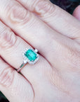 An emerald engagement ring with halo diamond on a hand with a natural background.