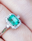 This inexpensive emerald engagement ring with halo diamond is on a finger.