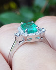 The underside view of an emerald engagement ring between fingers against a blurred natural background.